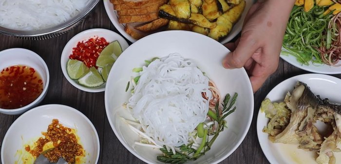 Assemble the noodles: Add vegetables (banana blossom, water spinach, water mimosa, lotus flower, rau răm) to the bottom of bowls. Add vermicelli noodles, broth, fish, and toppings (lemongrass, turmeric, chili).